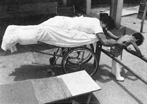 a man lying on belly on a wheel lying cart helping a child walk